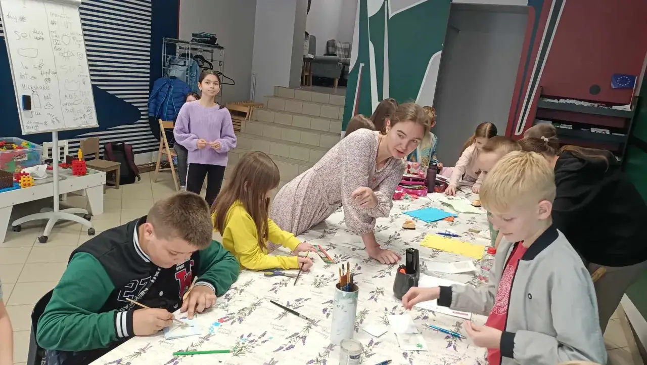 Dzieci biorą udział w zajęciach pozalekcyjnych, malując i rysując przy stole. Nauczycielka pomaga im w twórczej pracy.
