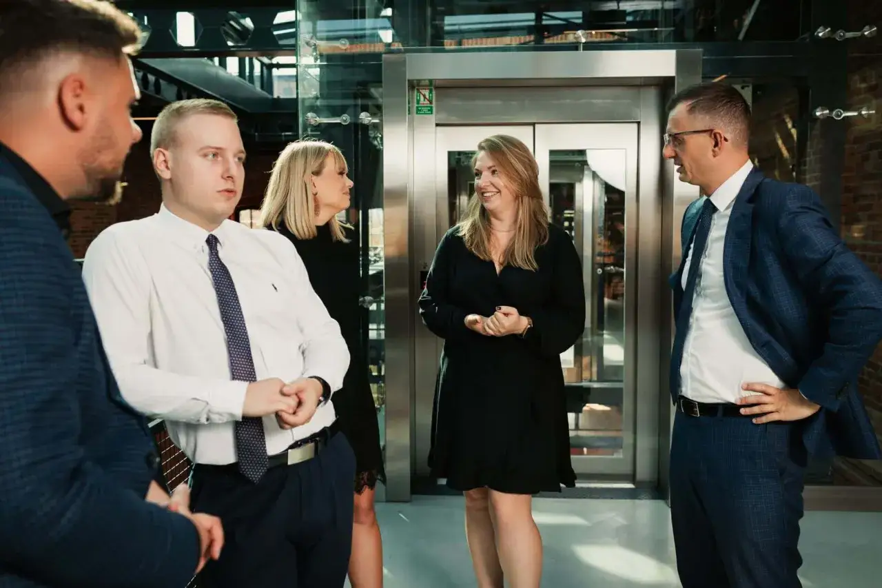 Grupa prawników w eleganckich strojach rozmawia przed windą w nowoczesnym biurze.
