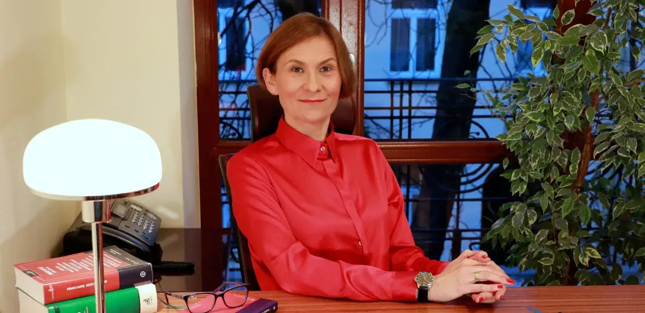 Kobieta w czerwonej koszuli siedzi przy biurku, obok lampka, książki i telefon.