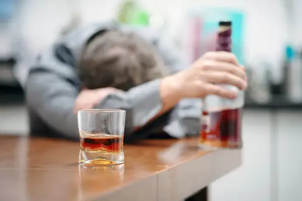 Ubezwłasnowolnienie alkoholika. Mężczyzna w garniturze leży na blacie, obok szklanka z whisky i butelka.