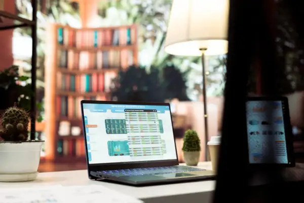 Laptop z danymi finansowymi, w tym wykresem i tabelami, prezentuje excel indeks. Obok stoi tablet, a w tle widać regały z książkami.