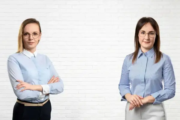 Dwie kobiety w koszulach i okularach na tle białej ceglanej ściany.