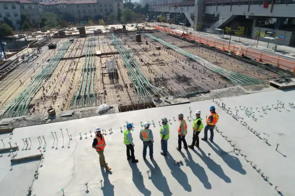 Grupa budowlańców w kaskach i kamizelkach omawia postęp prac na budowie. W tle widać zbrojenie i instalacje, zgodne z art. 62 prawa budowlanego.