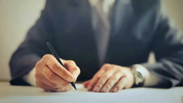 Dłoń w garniturze pisząca długopisem na białym papierze.