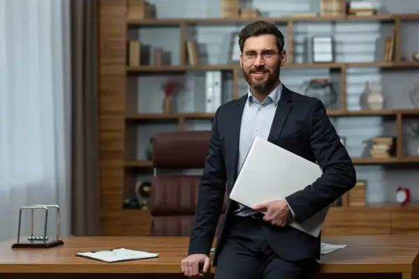 Uśmiechnięty dyrektor szkoły w garniturze, trzymający laptopa, stoi w swoim gabinecie.