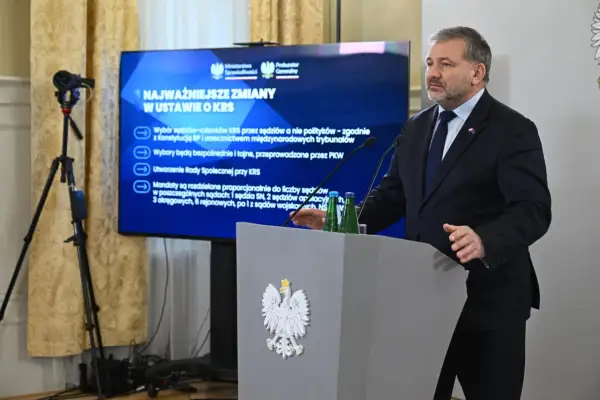 Mężczyzna przemawia przy mównicy z godłem Polski. Na ekranie za nim widnieje napis "Najważniejsze zmiany w ustawie o KRS", omawiający reformę neo KRS.