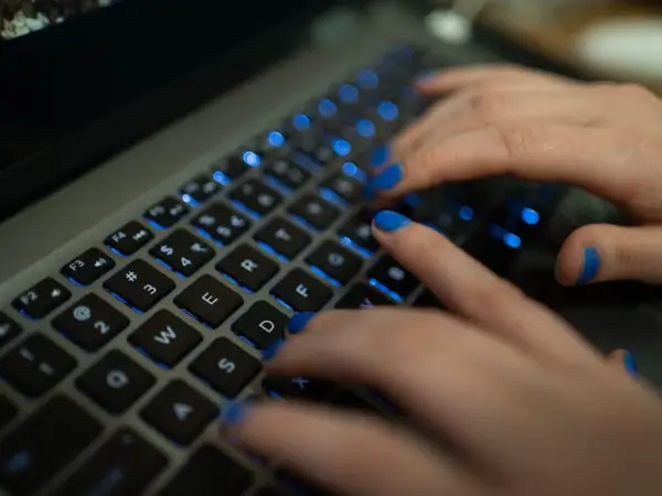 Dłonie z niebieskim lakierem na paznokciach piszą na podświetlanej klawiaturze MacBooka. Widać klawisze "twarda spacja mac".