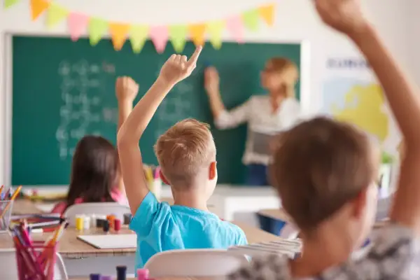 Uczniowie z podniesionymi rękami, gotowi do odpowiedzi. Wychowawca stoi przy tablicy, tłumacząc lekcję.
