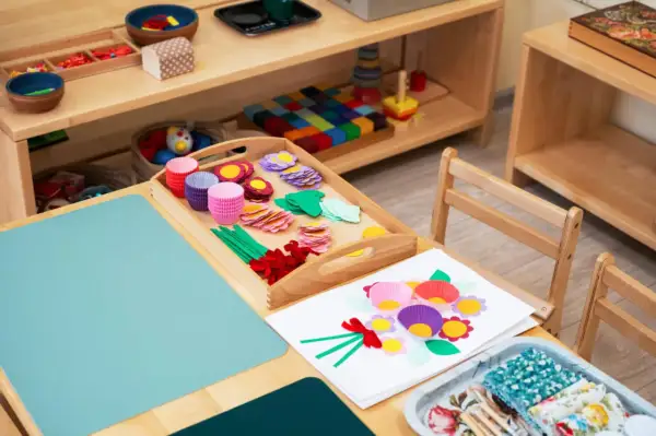 Zestaw do tworzenia kwiatów metodą Montessori: kolorowe papierowe foremki, listki, kółka i wstążki na drewnianej tacy.